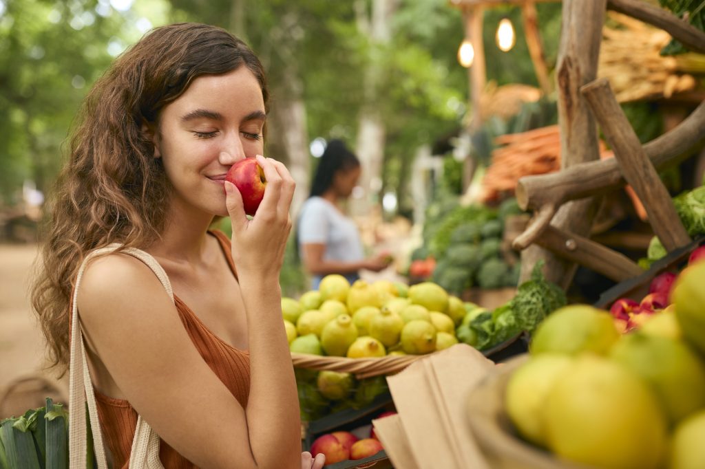 Woman smelling a fresh apple at a farmer’s market filled with fruits and vegetables